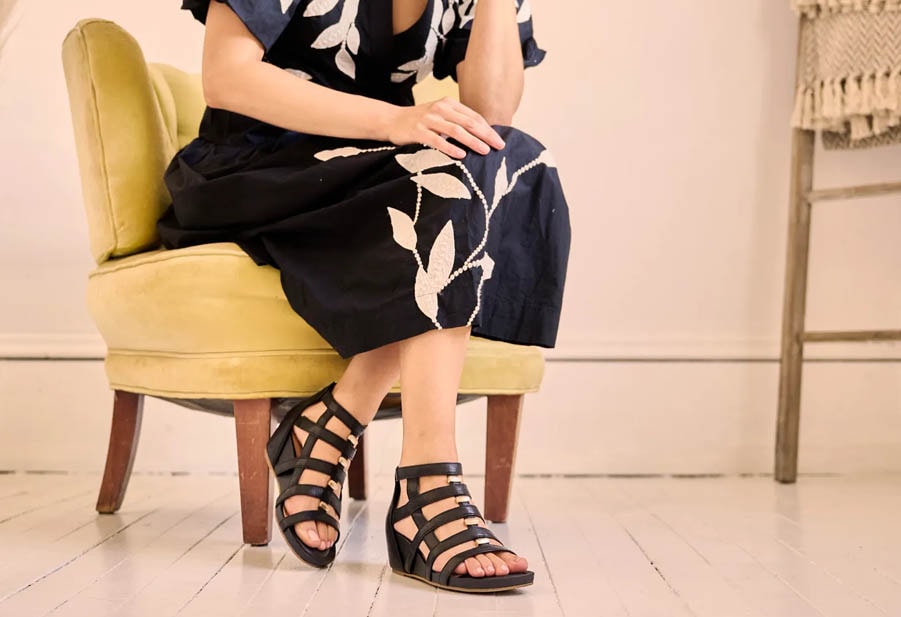Woman wearing a pair of Rio II wedge gladiator sandals in Black sitting on a yellow chair.